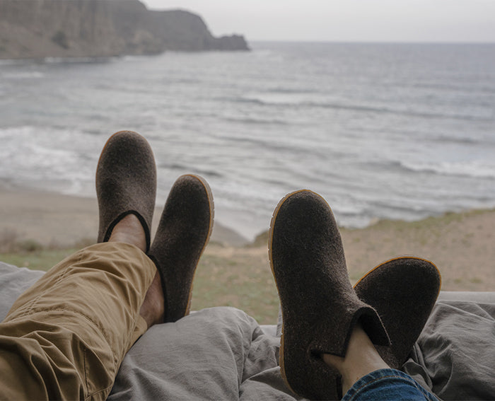 Two people relaxing with their feet up, looking out over a sandy beach and ocean waves under an overcast sky; both wearing Chestnu. brown slippers with honey rubber sole.