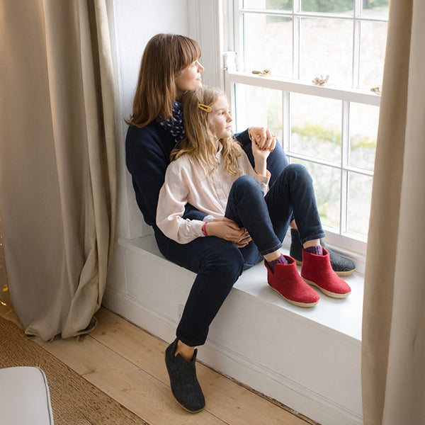 Two people are sitting by a window indoors, wearing glerups boos in Red and Charcoal grey colors.