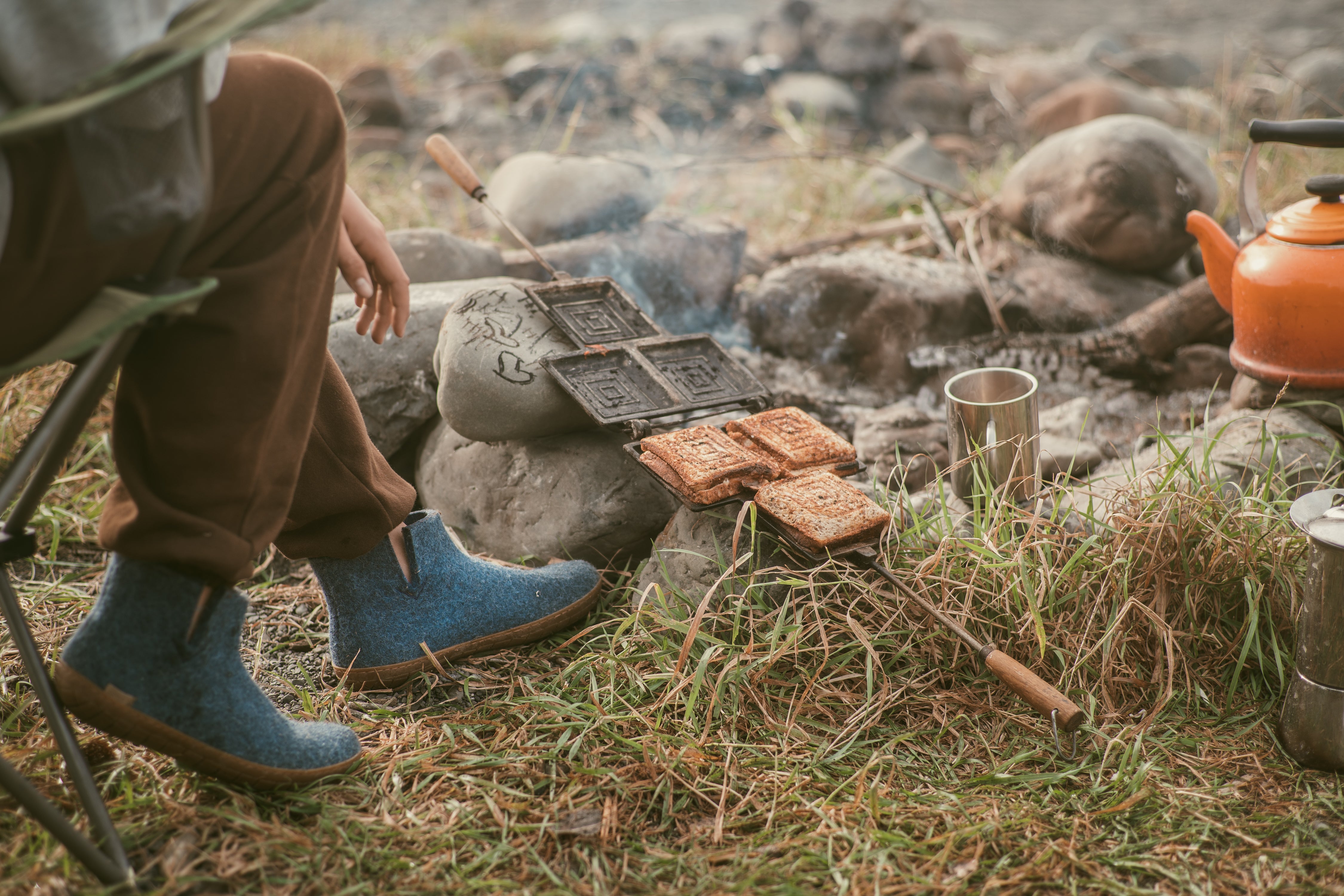 An outdoor, low-angle shot of a person sitting in a camping chair by a campfire, making toasted sandwiches in a cast-iron sandwich maker resting over the rocks, while wearing Denim, blue glerups boots with honey rubber soles.