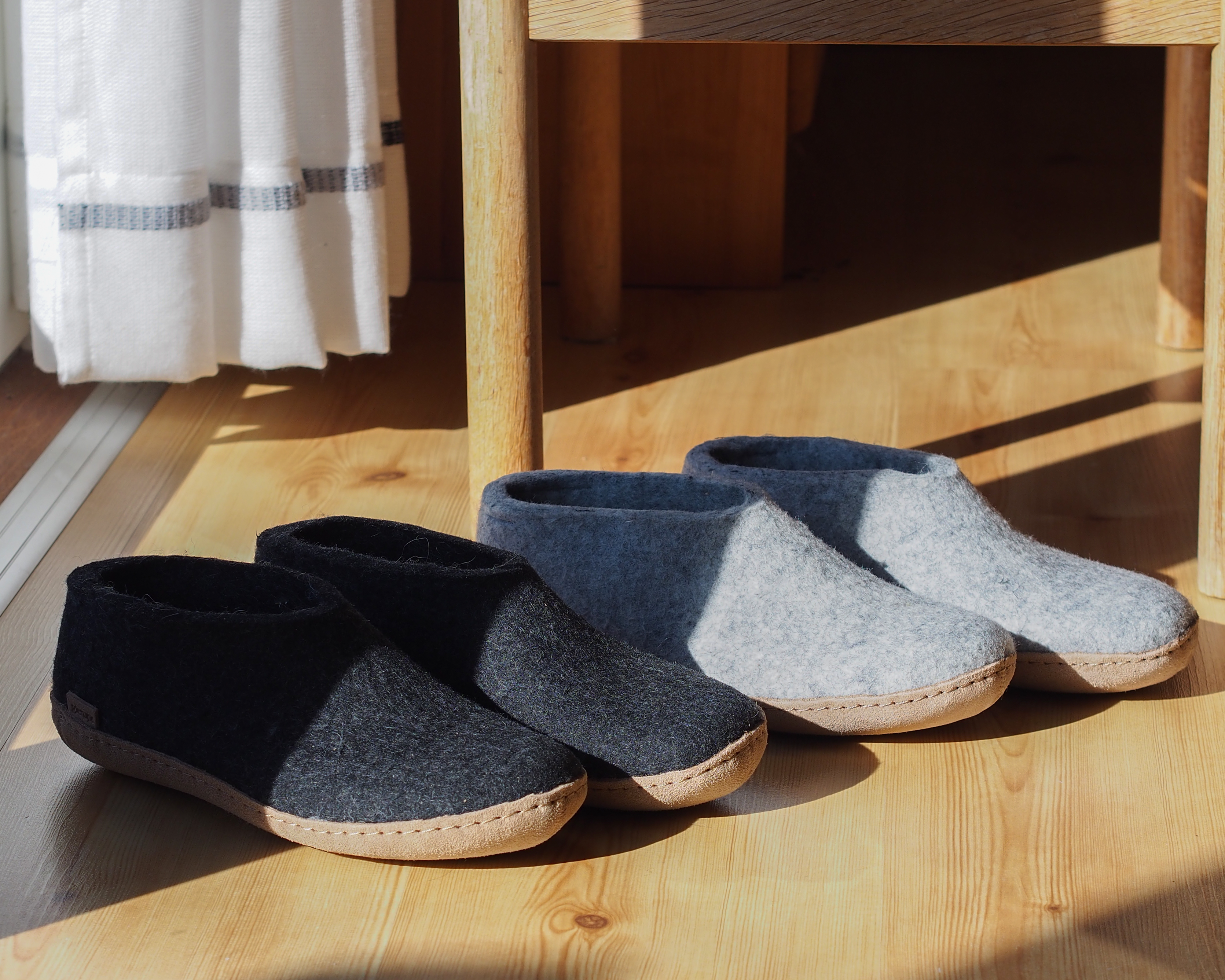 An indoor, high-angle shot of two pairs of felted glerups slip-on slippers sitting in a patch of bright morning sunlight on a light-colored hardwood floor near a wooden chair and a white striped curtain; one pair is Charcoal, black and the other is Grey, and both have leather soles shoe.